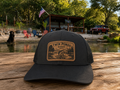 A black trucker hat with a brown leather patch reading 'Twin Ponds Ranch' sits on a wooden dock with a lake, trees, and outdoor cabin in the background.